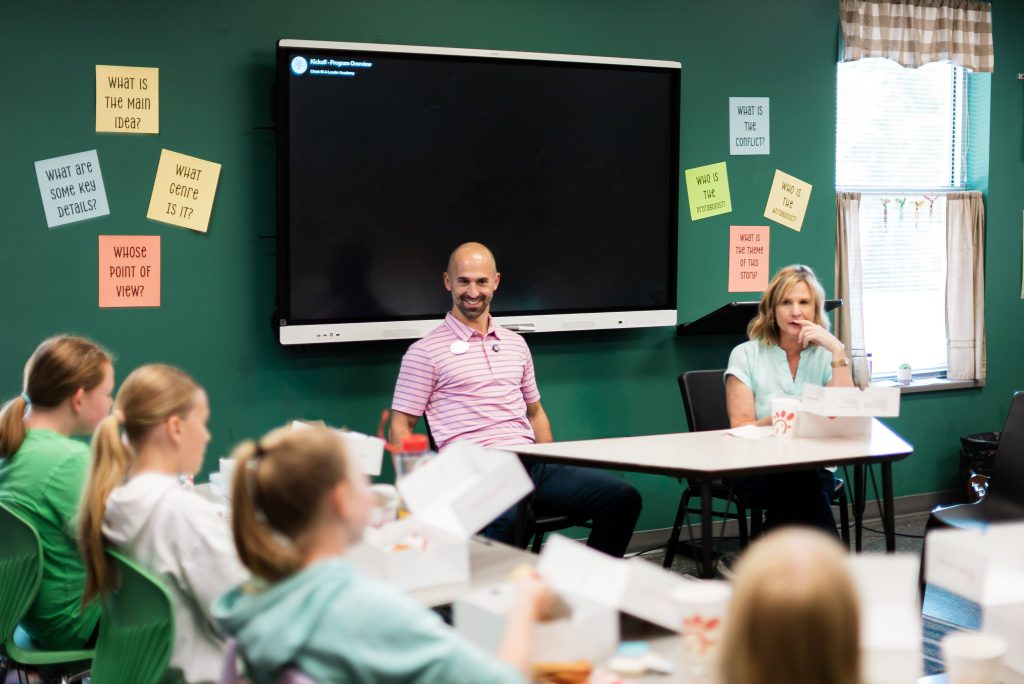 Corey Seitz, in a pink striped polo, smiles while speaking with students at the front of a classroom.