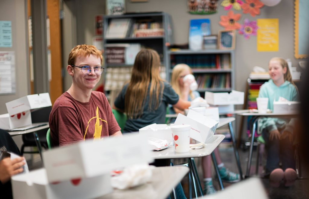 A smiling student in glasses sits at his desk while classmates eat lunch in the background.