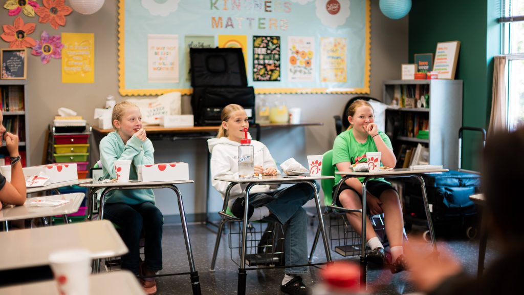 Three middle school students sit at desks with Chick-fil-A lunches, smiling and listening in a colorful classroom.